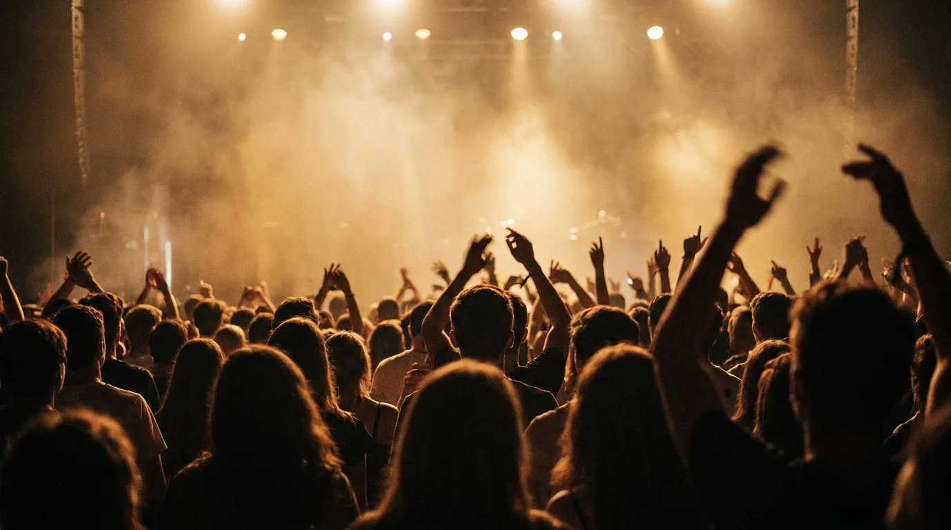 A blurred crowd of young concertgoers raising their hands toward a stage under warm spotlights