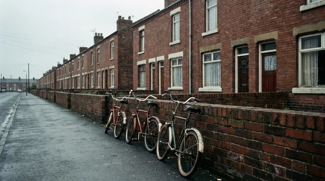 A row of identical red brick terraced houses on a quiet British street with three bicycles leaning against a low garden wall